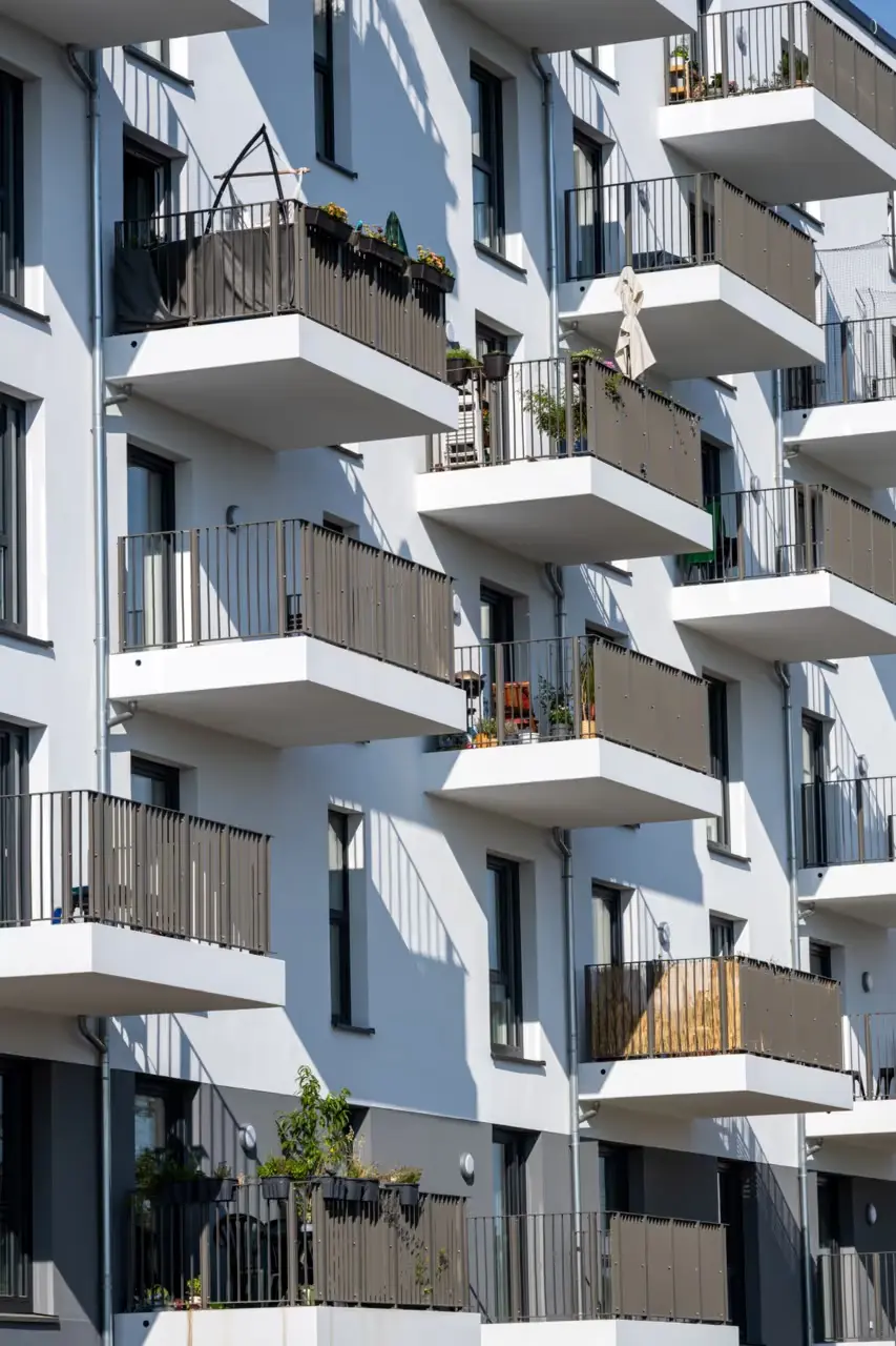 LUBA Immobilien UG – Immobilien – Moderne Fassade eines Mehrfamilienhauses mit mehreren Balkonen, die mit Metallgeländern ausgestattet sind und teilweise mit Pflanzen und Gartenmöbeln dekoriert sind.