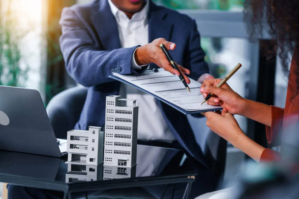 LUBA Immobilien UG - Immobilien - Zwei Personen in einer Besprechung, eine hält ein Klemmbrett und die andere unterzeichnet ein Dokument, auf dem Tisch stehen ein Laptop und Architekturmodelle.