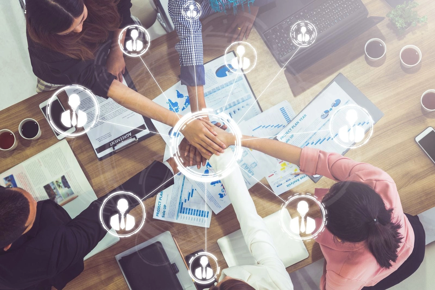 LUBA Immobilien UG - Immobilien - Draufsicht auf vier Geschäftspartner, die sich an einem Tisch mit Laptops, Dokumenten und Kaffeetassen die Hände reichen und so eine Geste der Teamarbeit zeigen.
