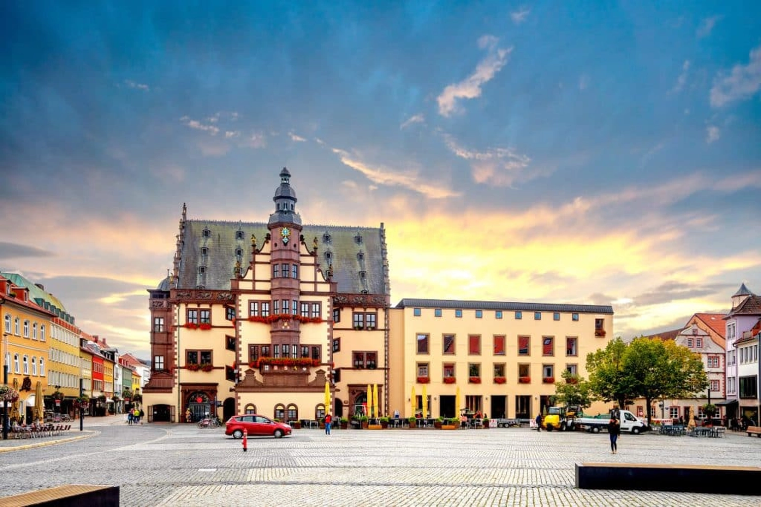 LUBA Immobilien UG - Immobilien - Historisches Gebäude mit kunstvoller Architektur und Uhrturm auf einem gepflasterten Marktplatz bei Sonnenuntergang.