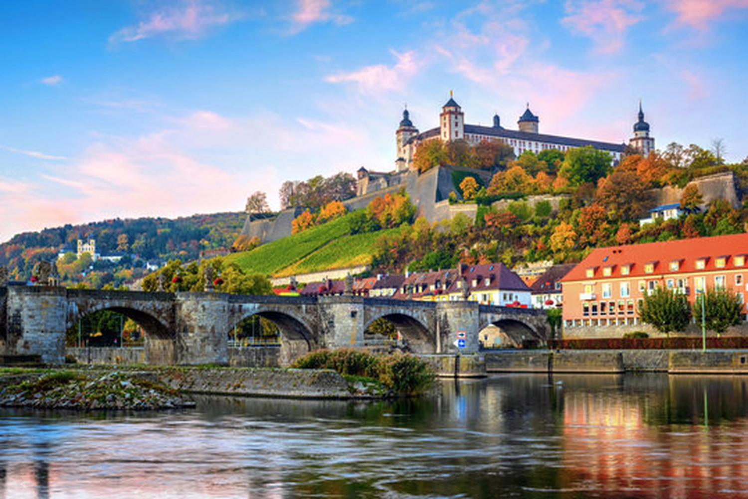 LUBA Immobilien UG – Immobilien – Steinbrücke über einen Fluss mit einer Burg am Hang und bunten Herbstbäumen im Hintergrund unter blauem Himmel mit rosa Wolken