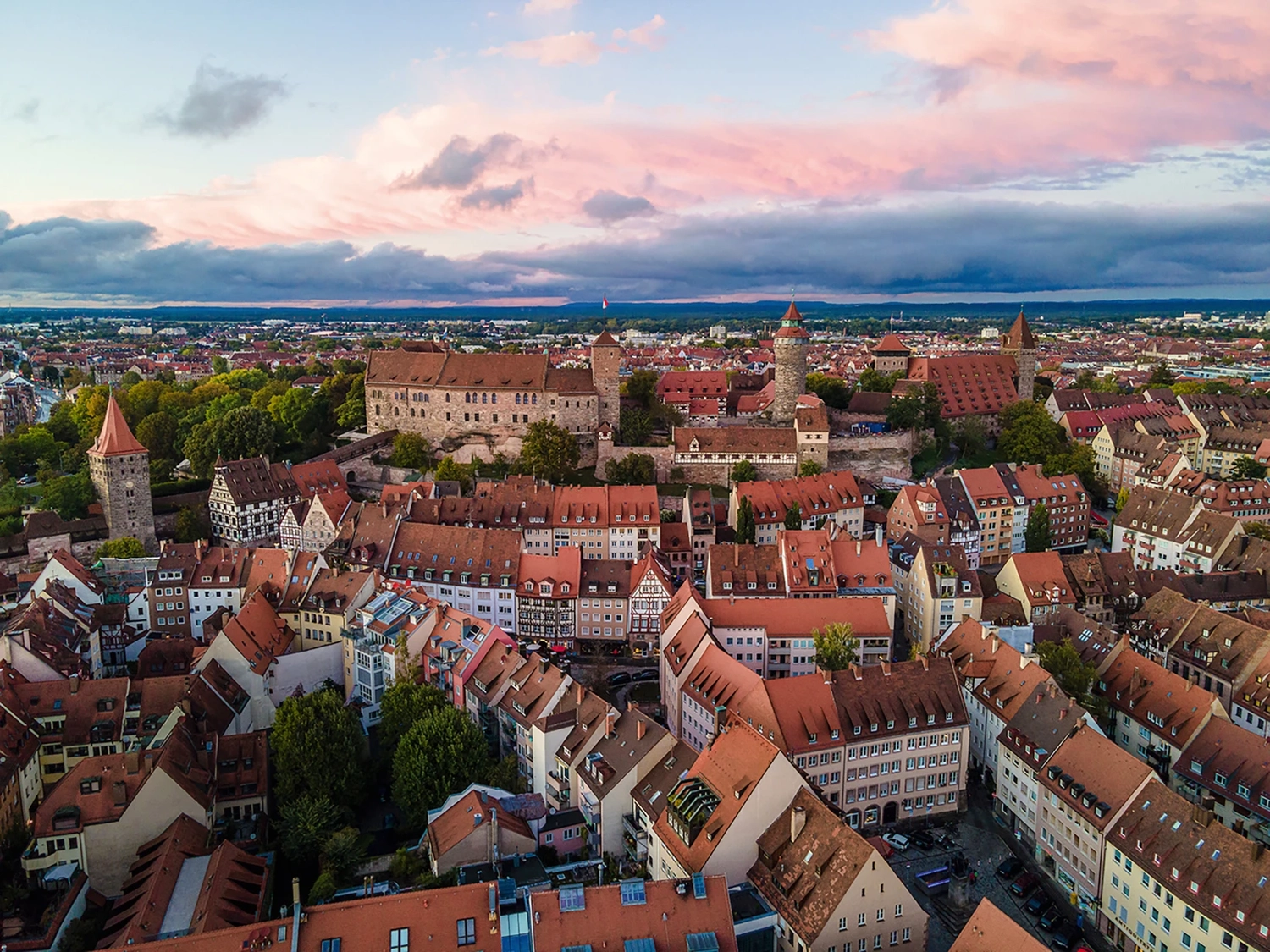 LUBA Immobilien UG – Immobilien – Luftaufnahme einer historischen europäischen Stadt mit roten Ziegeldächern, imposanten steinernen Burgmauern und Türmen unter einem rosa-blauen Sonnenuntergangshimmel