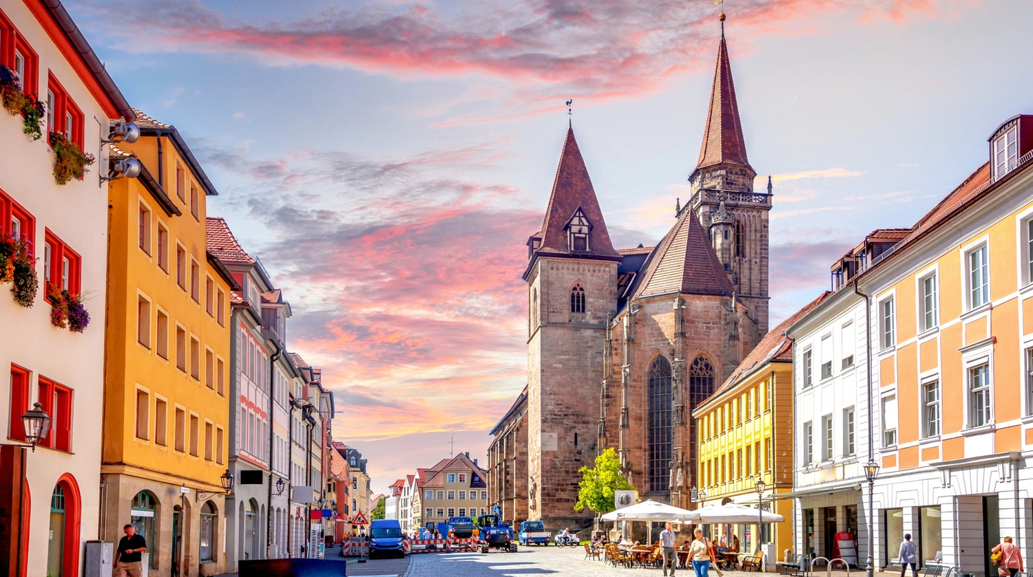 LUBA Immobilien UG – Immobilien – Historische europäische Straße mit farbenfrohen Gebäuden, Straßencafés und einer großen Steinkirche mit spitzen Türmen unter einem rosa-blauen Sonnenuntergangshimmel