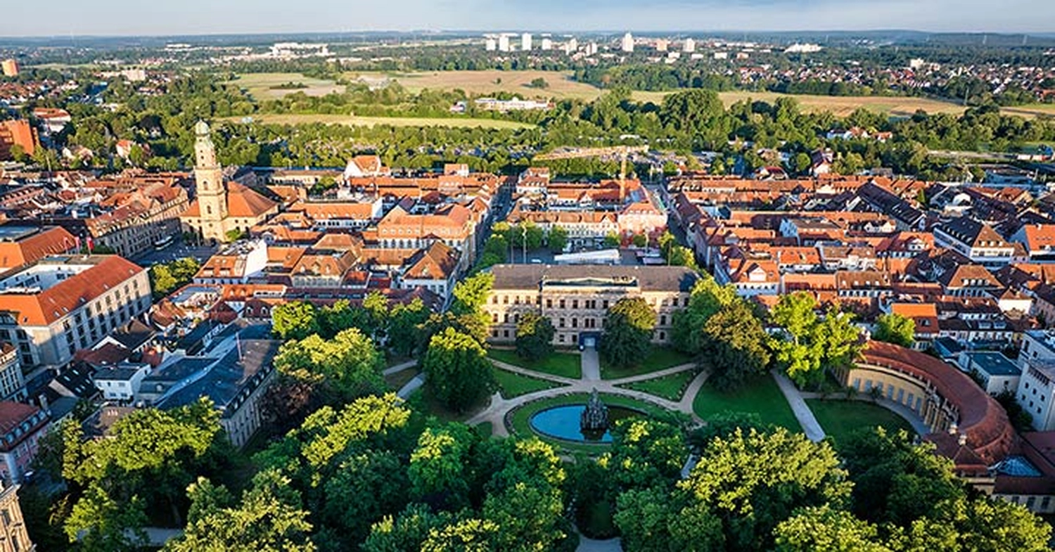 LUBA Immobilien UG - Immobilien - Luftaufnahme einer europäischen Stadt mit rotgedeckten Gebäuden, einem markanten Kirchturm, einem klassischen Gebäude an einem runden Teich und grünen Parks