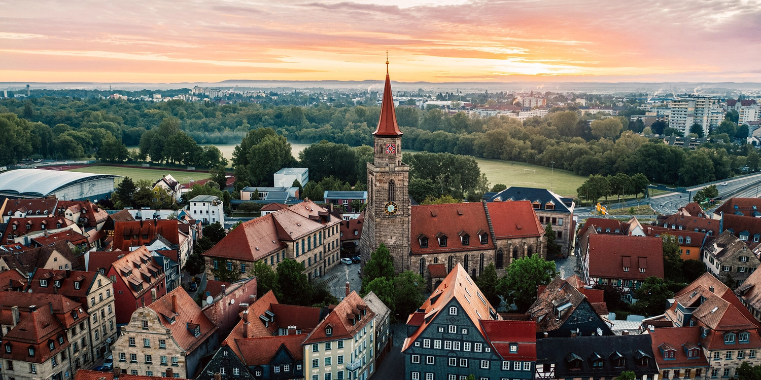 LUBA Immobilien UG – Immobilien – Luftaufnahme einer historischen Stadt mit rotgedeckten Gebäuden, einem markanten Kirchturm, grünen Bäumen und einem Sonnenuntergang