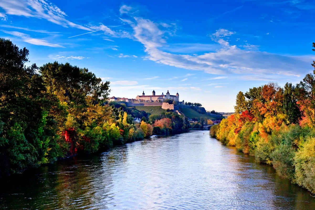LUBA Immobilien UG – Immobilien – Ein von Bäumen mit herbstlichem Laub gesäumter Fluss und ein historisches Schloss auf einem Hügel unter einem teilweise bewölkten blauen Himmel